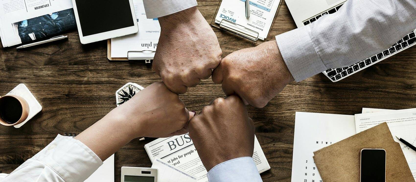 Vier geballte Hände über einen Arbeitstisch, die aneinanderstoßen und Teamwork auszudrücken. Auf dem Tisch liegen viele Unterlagen, Handy und Taschenrechner.